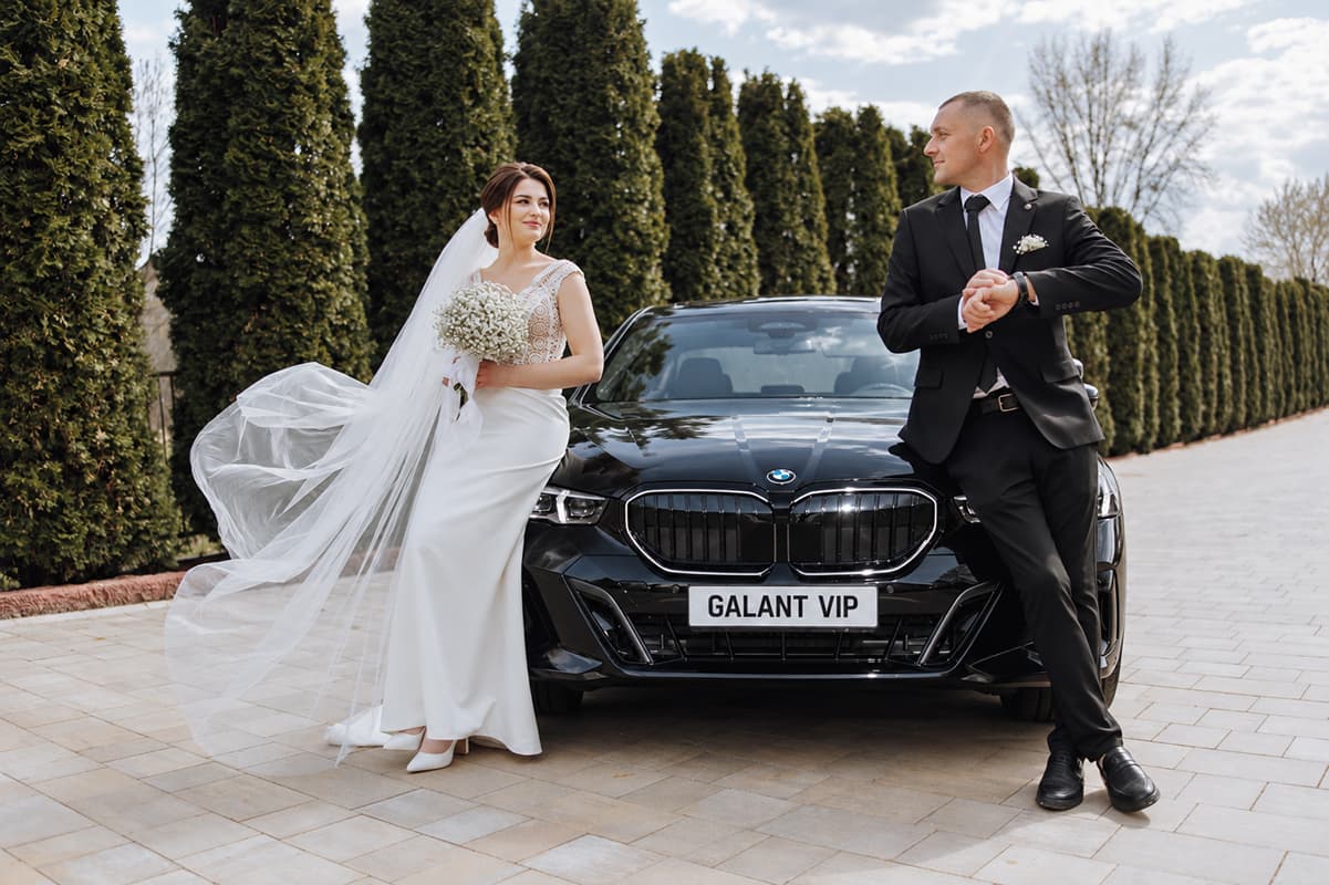 Mire și mireasă lângă un BMW negru Galant VIP, pe o alee mărginită de arbori.